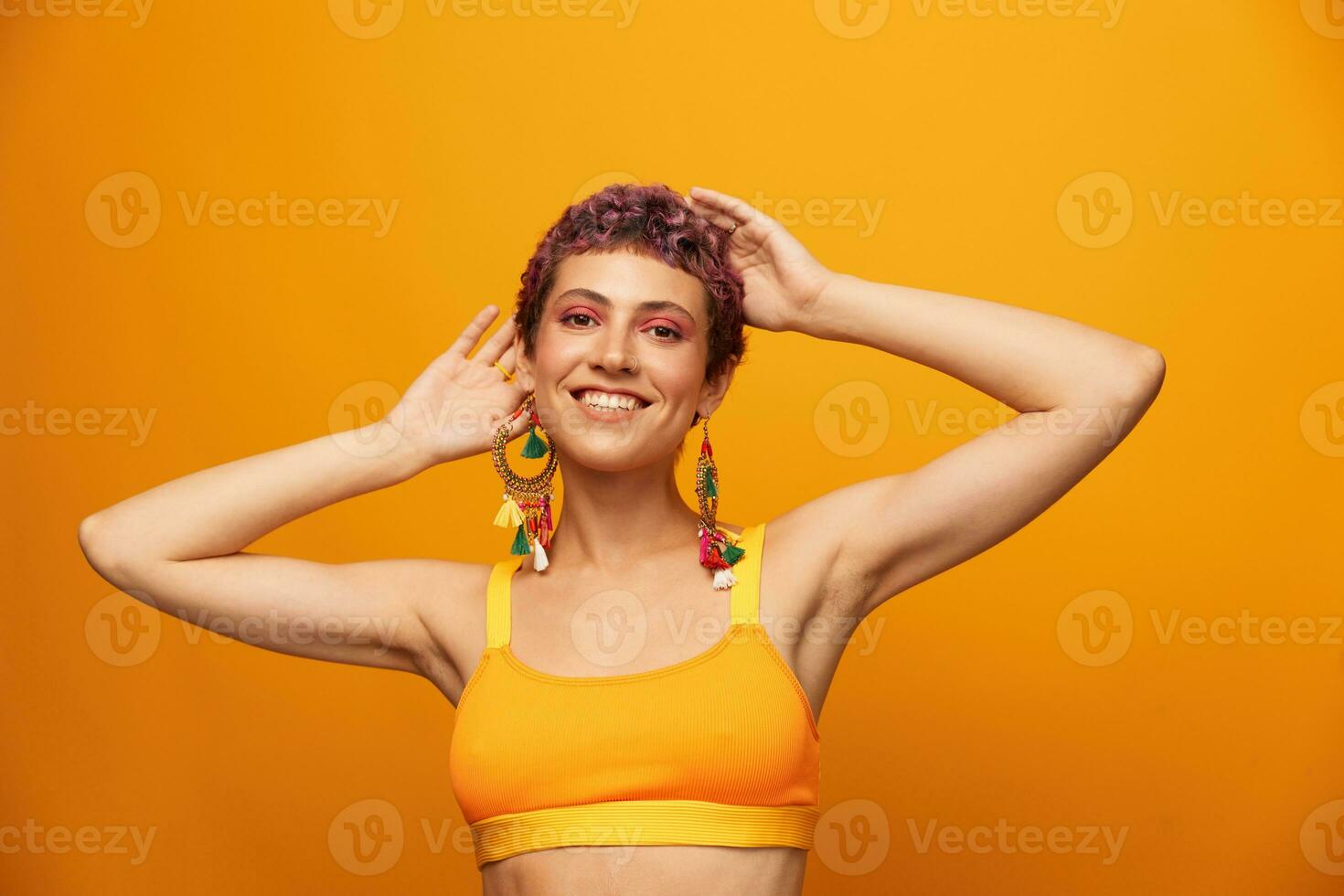 Portrait of a young woman with a short haircut and colored hair smiling and showing her tongue at the camera on an orange background with earrings accessories in the studio photo