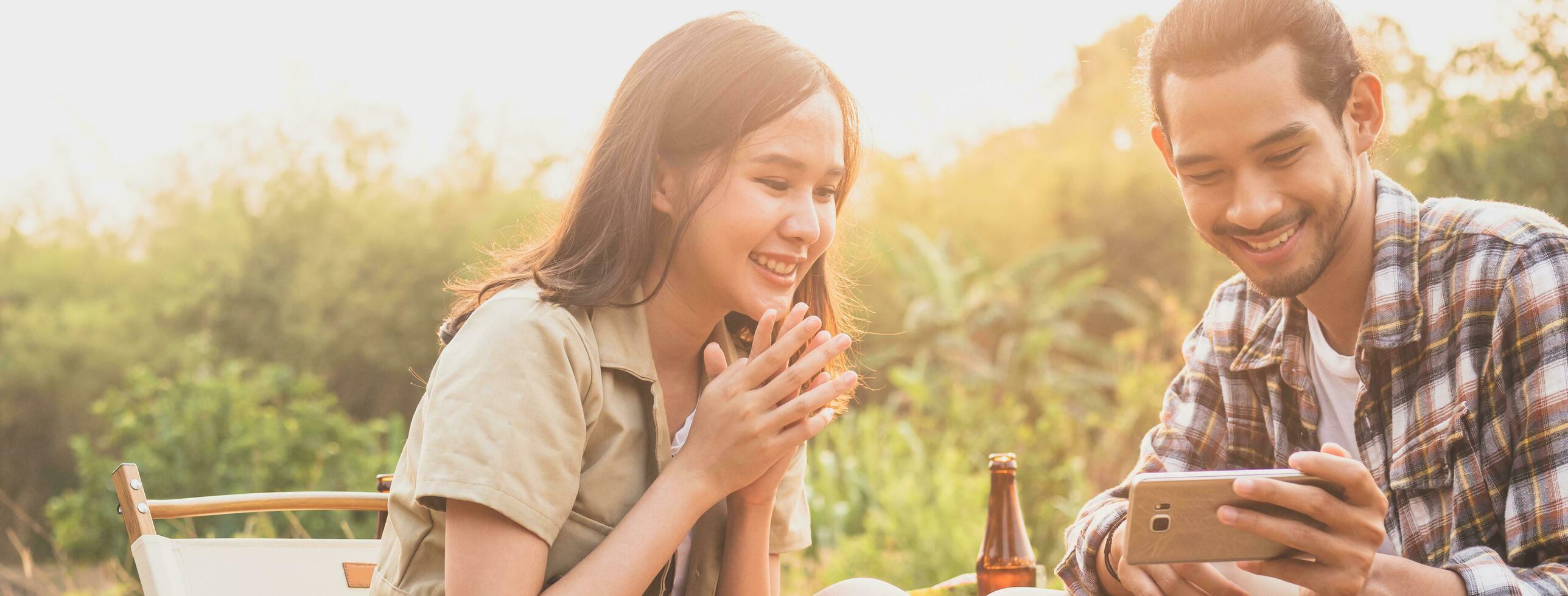 feliz, alegre asiático joven mujer, niña y hombre tenencia, utilizando teléfono inteligente sentado en silla fuera de carpa. aventuras Pareja amor cámping en bosque. eco actividad, estilo de vida naturaleza en fiesta concepto. foto