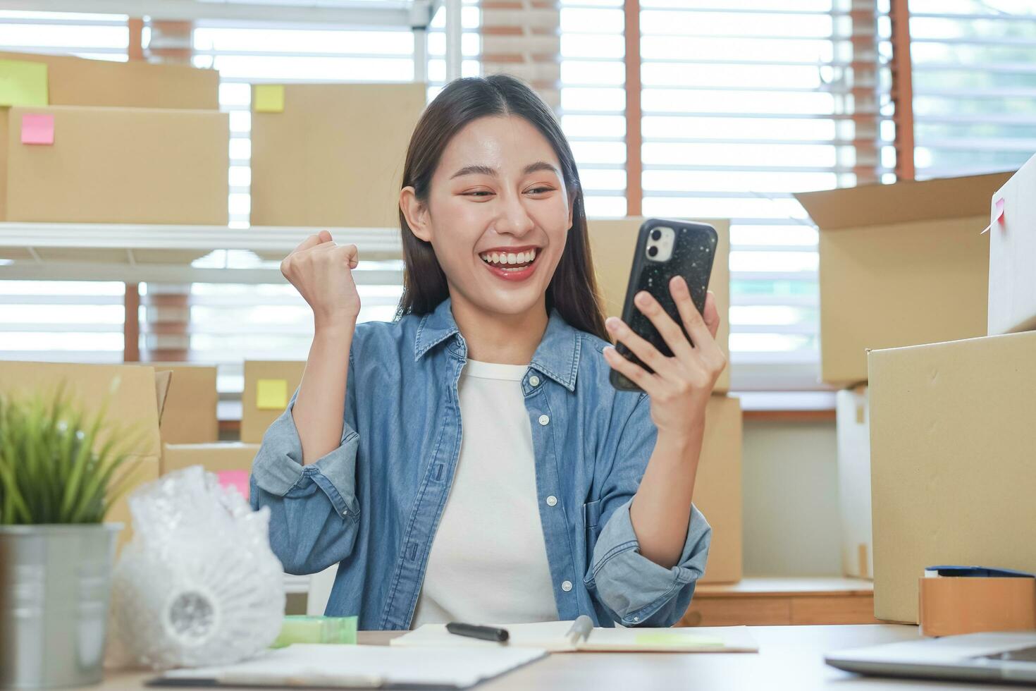 alegre pequeño negocio empresario pequeño, contento asiático joven mujer propietario sonrisa cuando recibir orden desde cliente en aplicación compras por teléfono inteligente, móvil. comerciante en línea, comercio electrónico trabajando a hogar oficina. foto