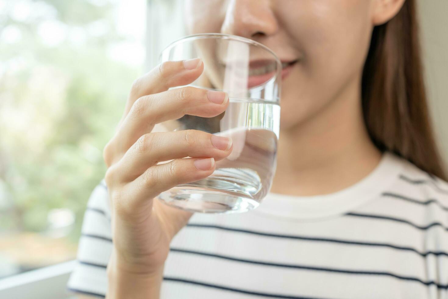 Happy beautiful, asian young woman, girl drinking, sip fresh glass of water for hydration of body, holding transparent glass in her hand, thirsty at home. Health care, healthy lifestyle concept. photo