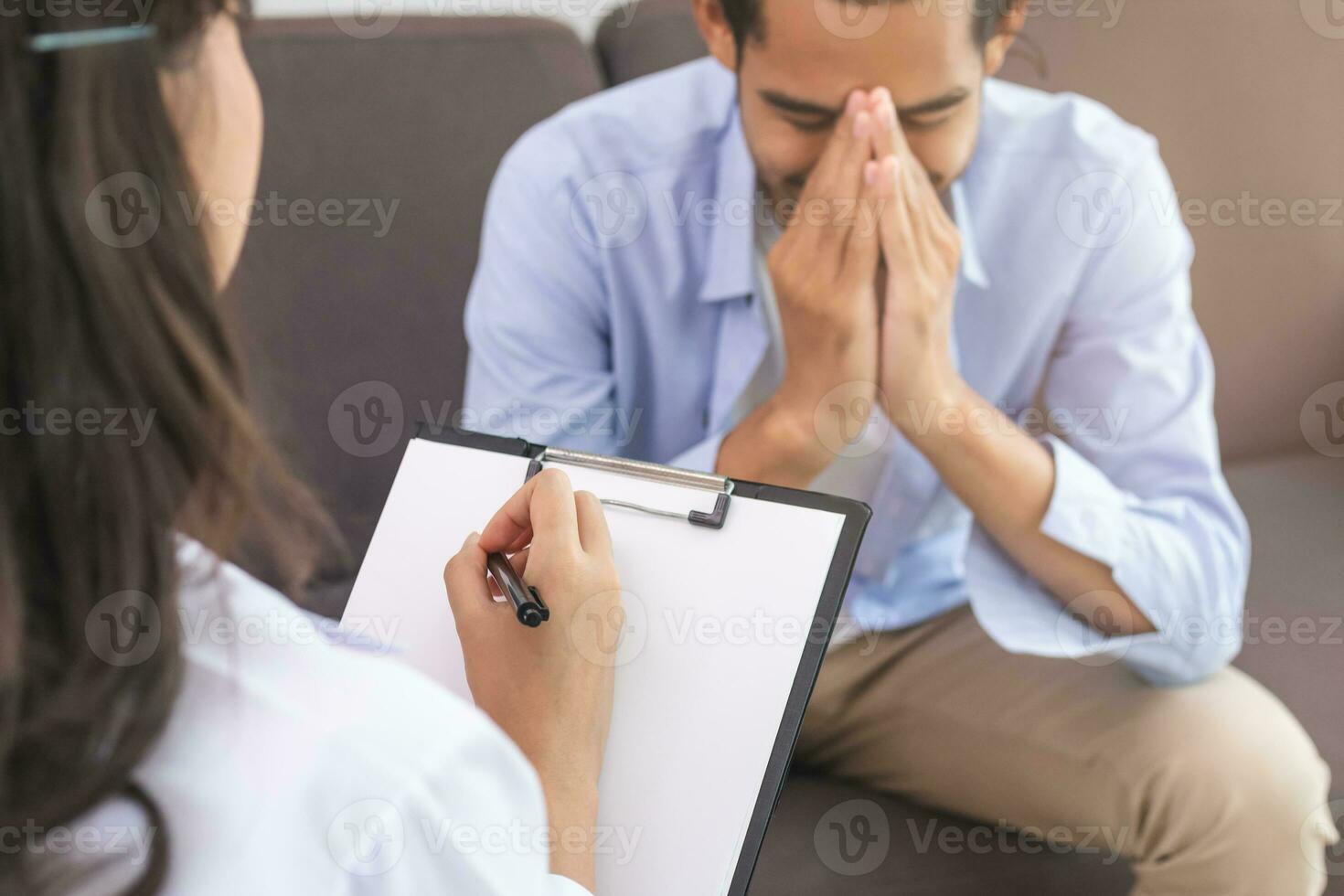 Asian young man, male has mental symptoms which must have been therapy and stress, sitting on couch to consult to psychologist during the session  taking notes to find out how to treat the therapist. photo