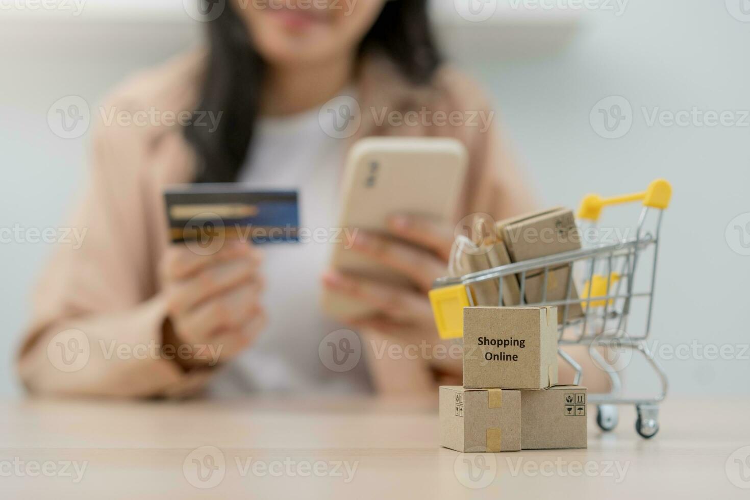 Cardboard box parcel in a trolley, asian young woman using mobile phone with shopping service on market place, website direct buy goods from seller over internet. Online shopping at home, delivery. photo