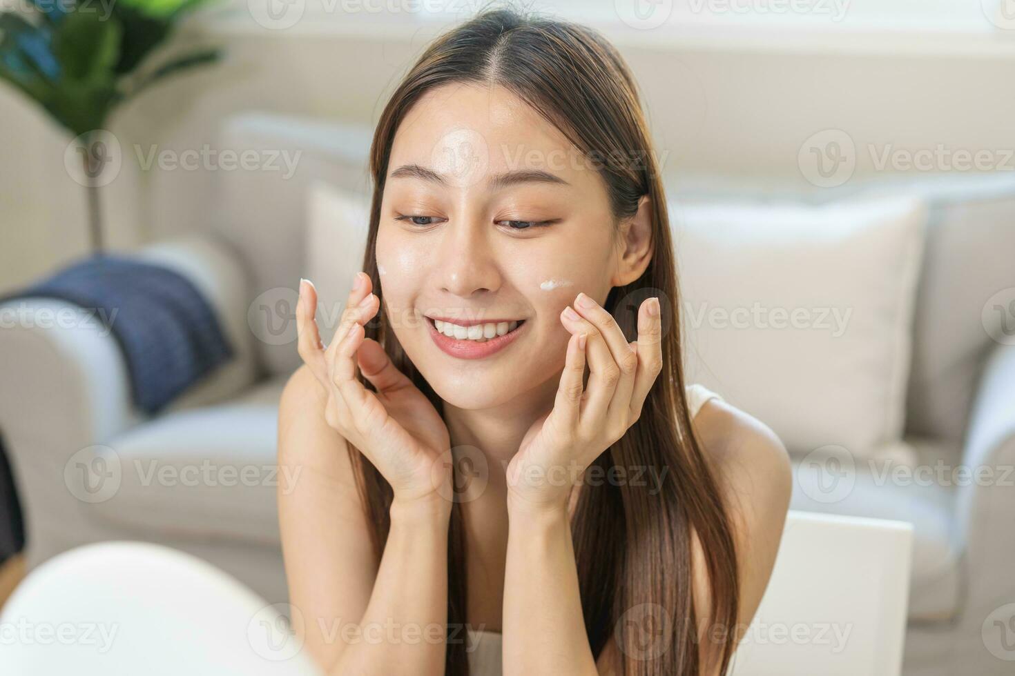 Fresco sano piel, hermosa sonrisa de asiático joven mujer, niña mirando a espejo, aplicando hidratante en su rostro, poniendo crema tratamiento antes de maquillaje cosmético rutina a hogar. facial belleza. foto