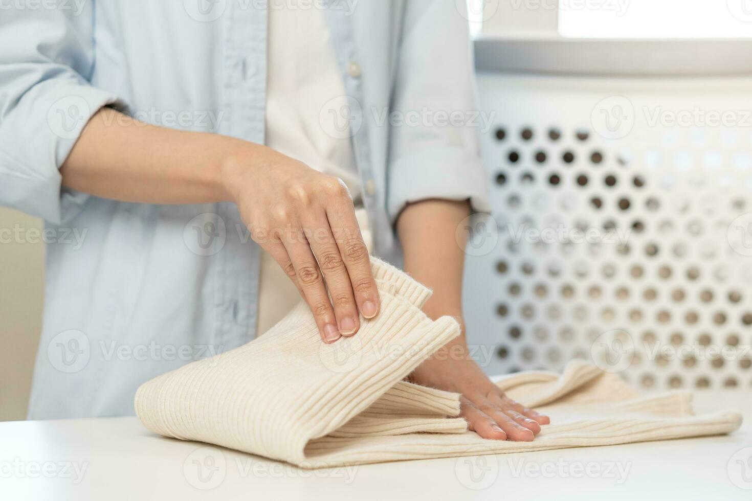 Housewife, asian young woman hand in many folding freshly shirts, sweaters or dress on desk, table after washing clean clothes and drying, making household working in room at home. Laundry and maid. photo