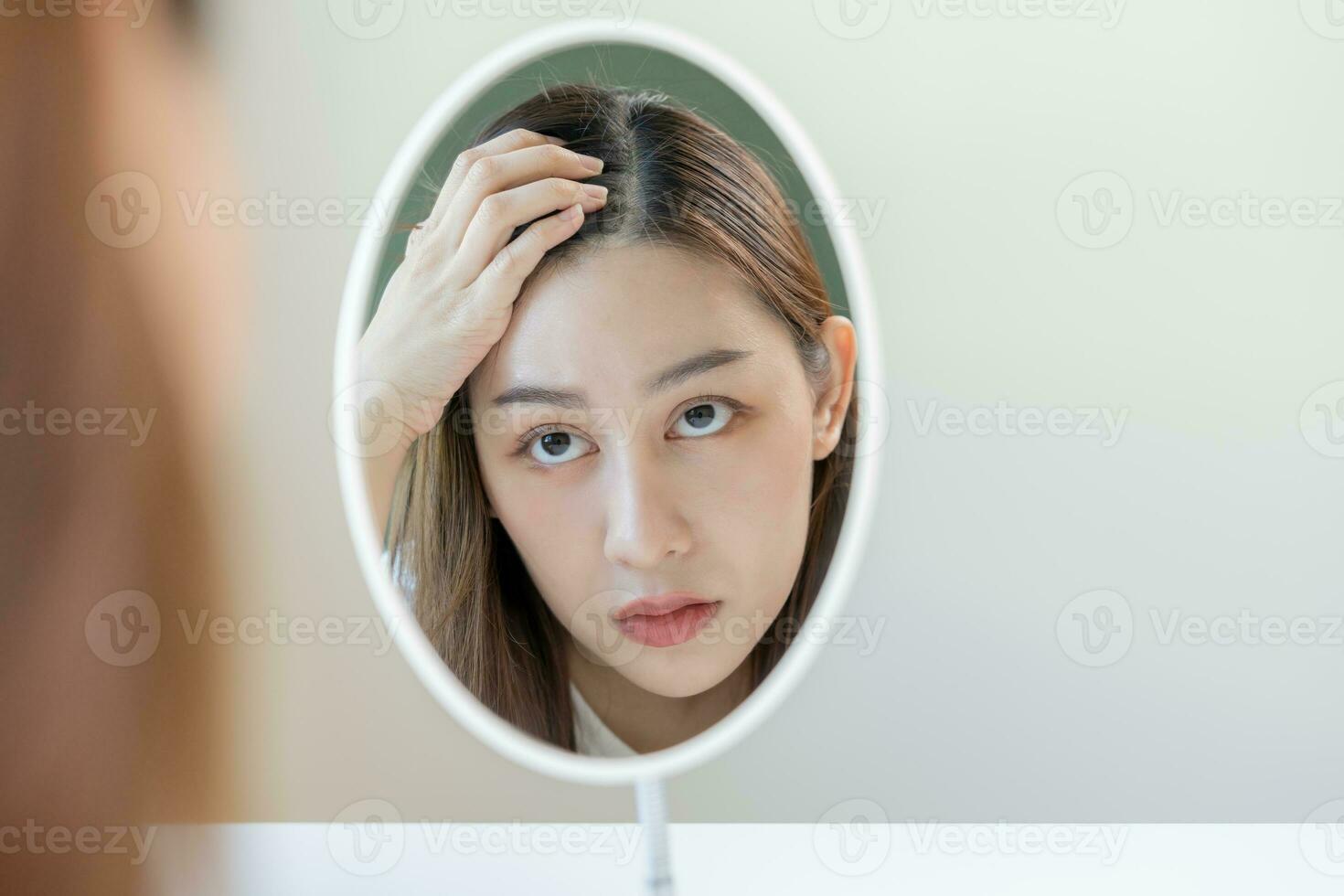 Damaged Hair, face serious asian young woman worry looking at scalp in mirror, hand in break into front hair loss, thin problem symptom at home. Health care shampoo beauty, isolated on background. photo