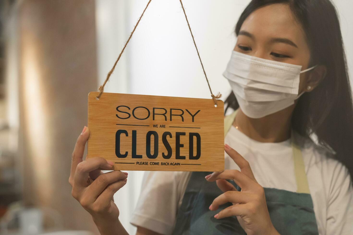 Asian young owner retail,coffee shop woman turning sign board to closed with wearing face mask,protection to pandemic of coronavirus.Close store,restaurant or pub due to lockdown, quarantine of covid. photo