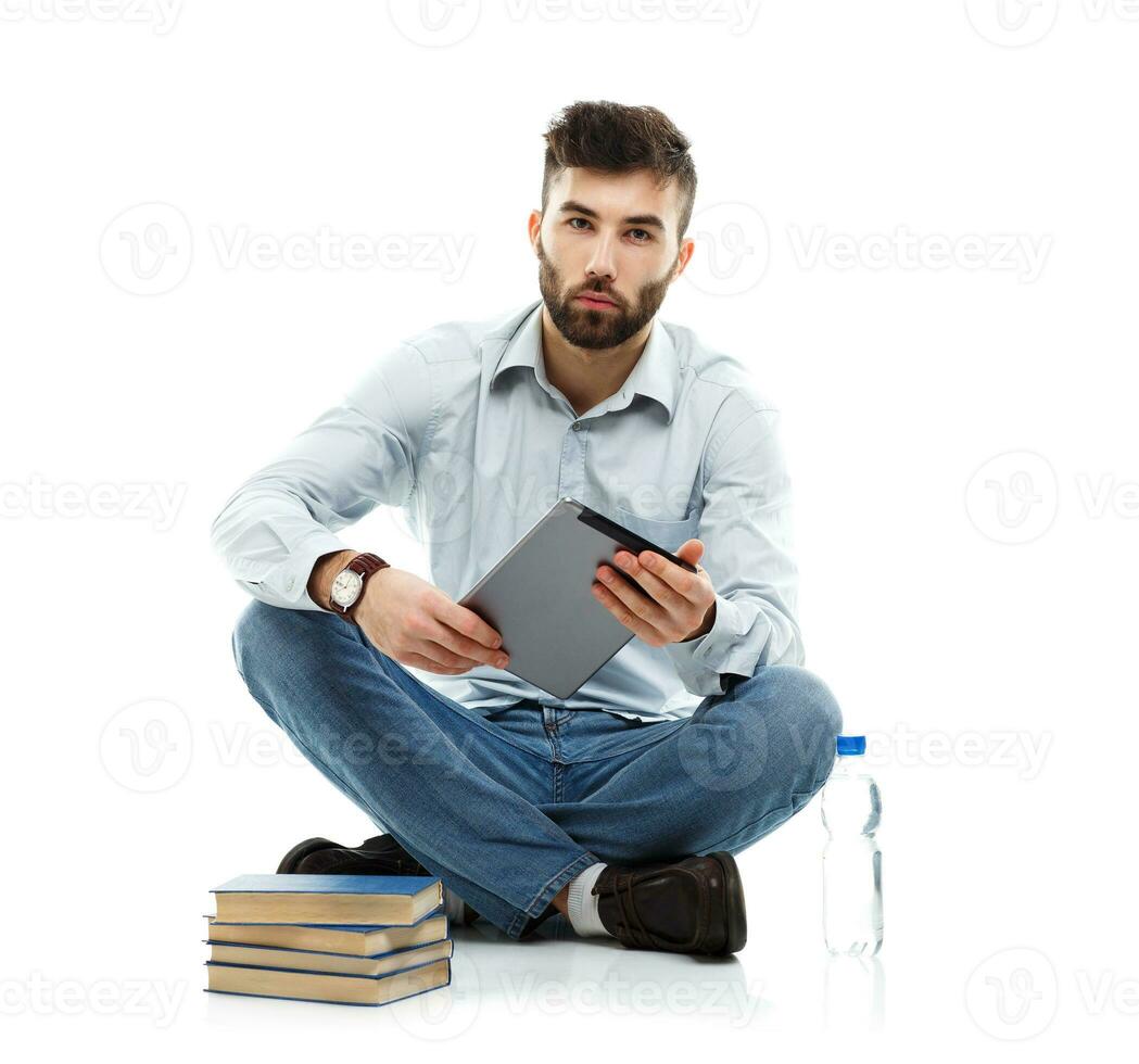 Young bearded smiling man holding a tablet with books and a bottle of water sitting on a white photo