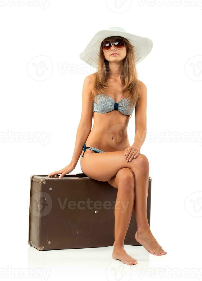 Full length portrait of a beautiful young woman posing in a bikini sitting on a suitcase on a white photo