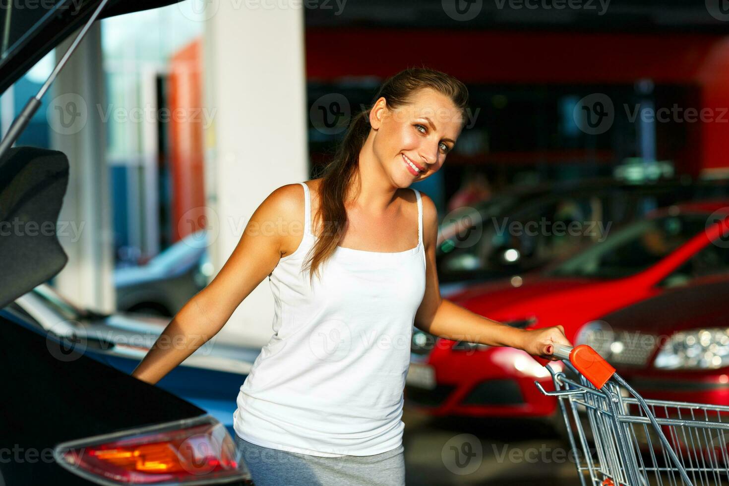 Young woman shifts the purchase from shopping cart in the trunk of a car on the parking photo