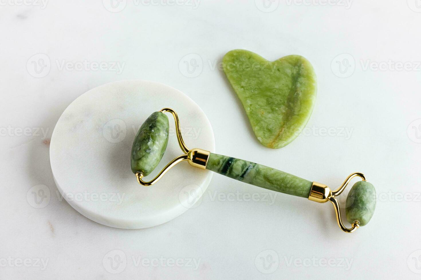 Green jade roller and gua sha stone massager for face on white marble. photo