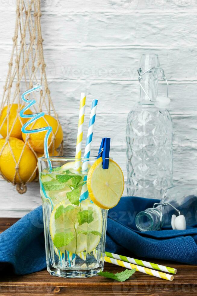 Lemonade with mint in glass, lemons in grid bag and bottles on wooden table. photo