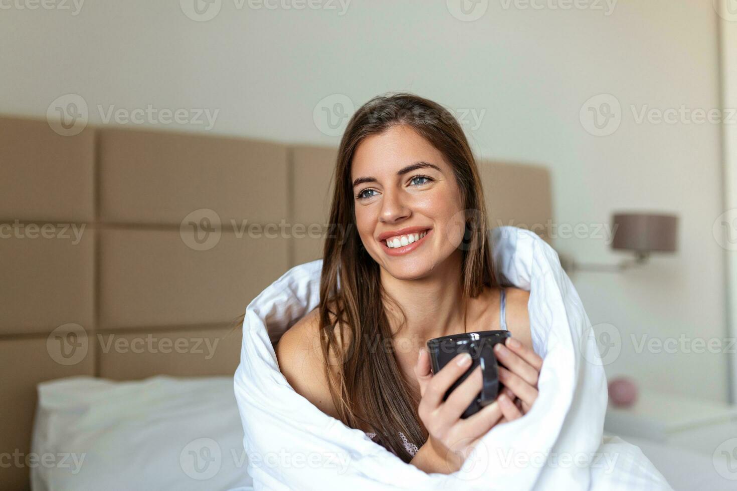 Beautiful young woman holding a cup of coffee while lying on bed. Morning concept. young female smiling and looking away while resting on bed and drinking hot beverage in morning at home photo
