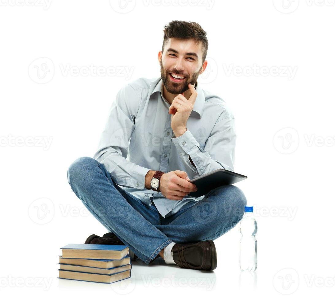 Young bearded smiling man holding a tablet with books and a bottle of water sitting on a white photo