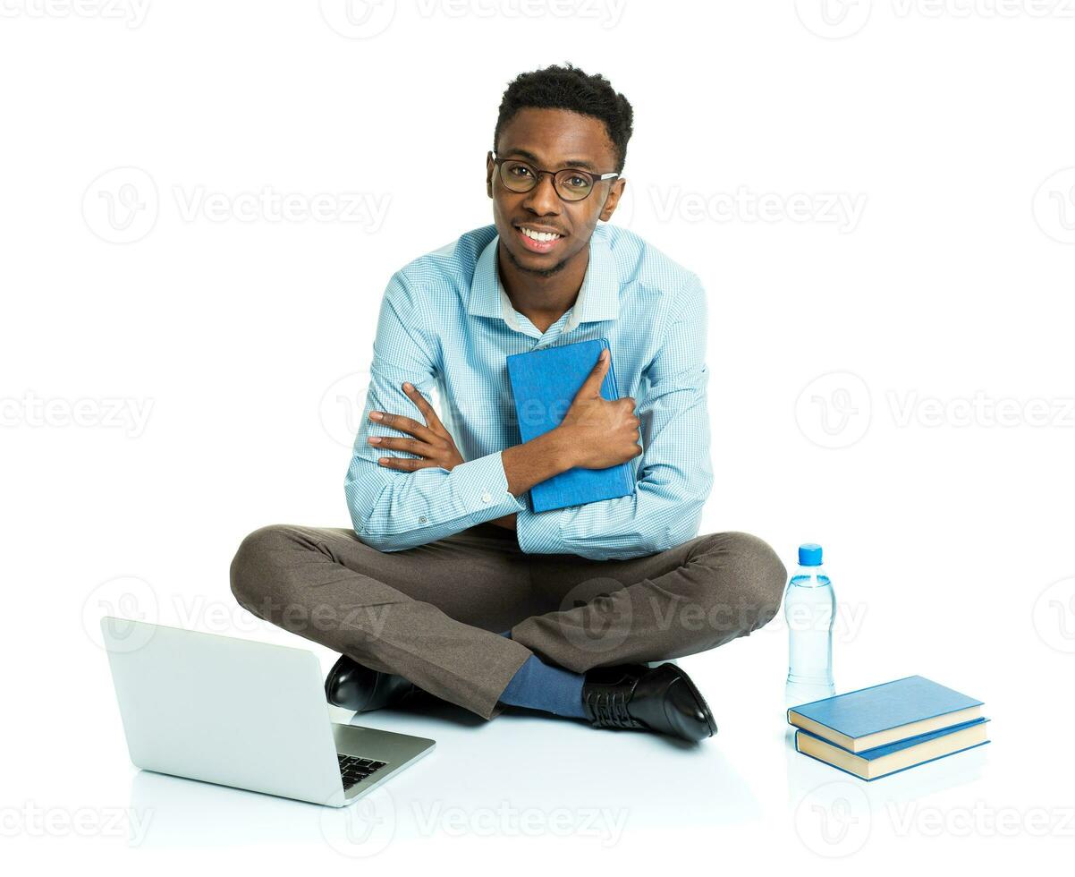 Happy african american college student with laptop, books and bottle of water sitting on white photo