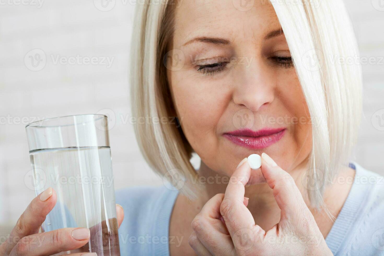 Doubtful sick ill middle woman holding pill and glass of water taking painkiller medicine drugs to relieve headache pain, worried about side effects of antidepressant or emergency contraceptive meds photo