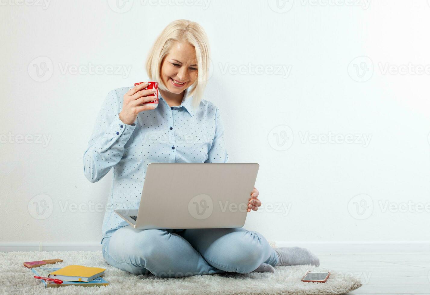 Happy middle aged woman sitting relaxed on the floor using laptop for entertainment. The concept of leisure and work with a cup of coffee at home. photo