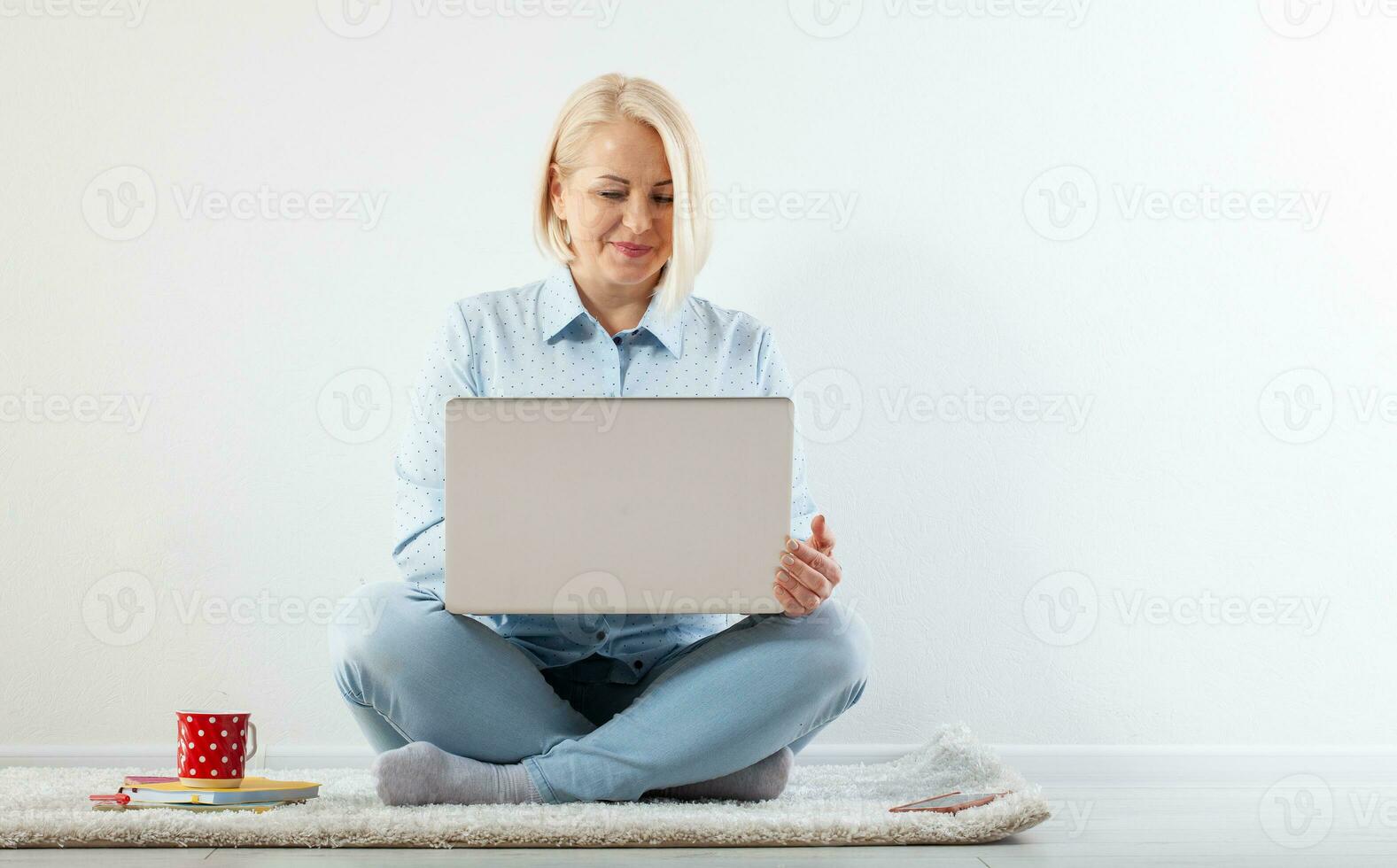 Happy middle aged woman sitting relaxed on the floor using laptop for entertainment. The concept of leisure and work with a cup of coffee at home. photo