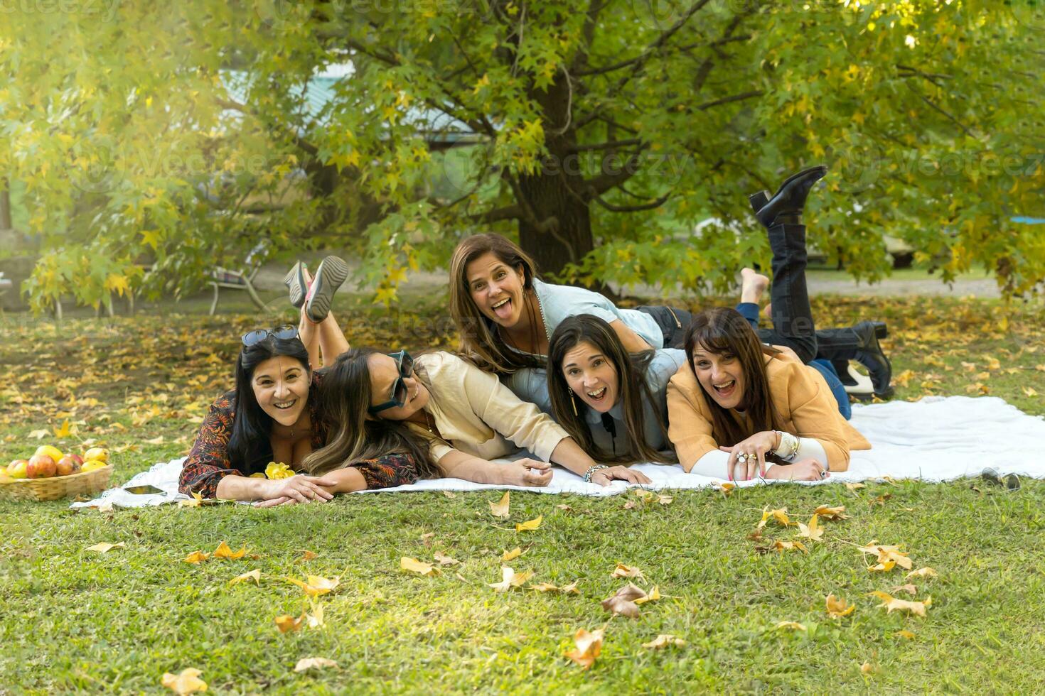 Group of friends having fun together lying on the grass while taking a self-portrait - Happy middle-aged women taking a group photo at a picnic at a campsite - Friendship and lifestyle concept