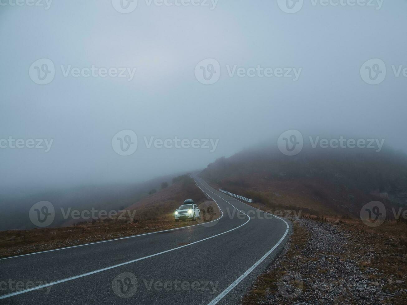 A Rover car SUV with fog lights turned on is parked next to a scenic route road in a foggy autumn landscape. Luggage trunk box mountain on car roof rack. photo