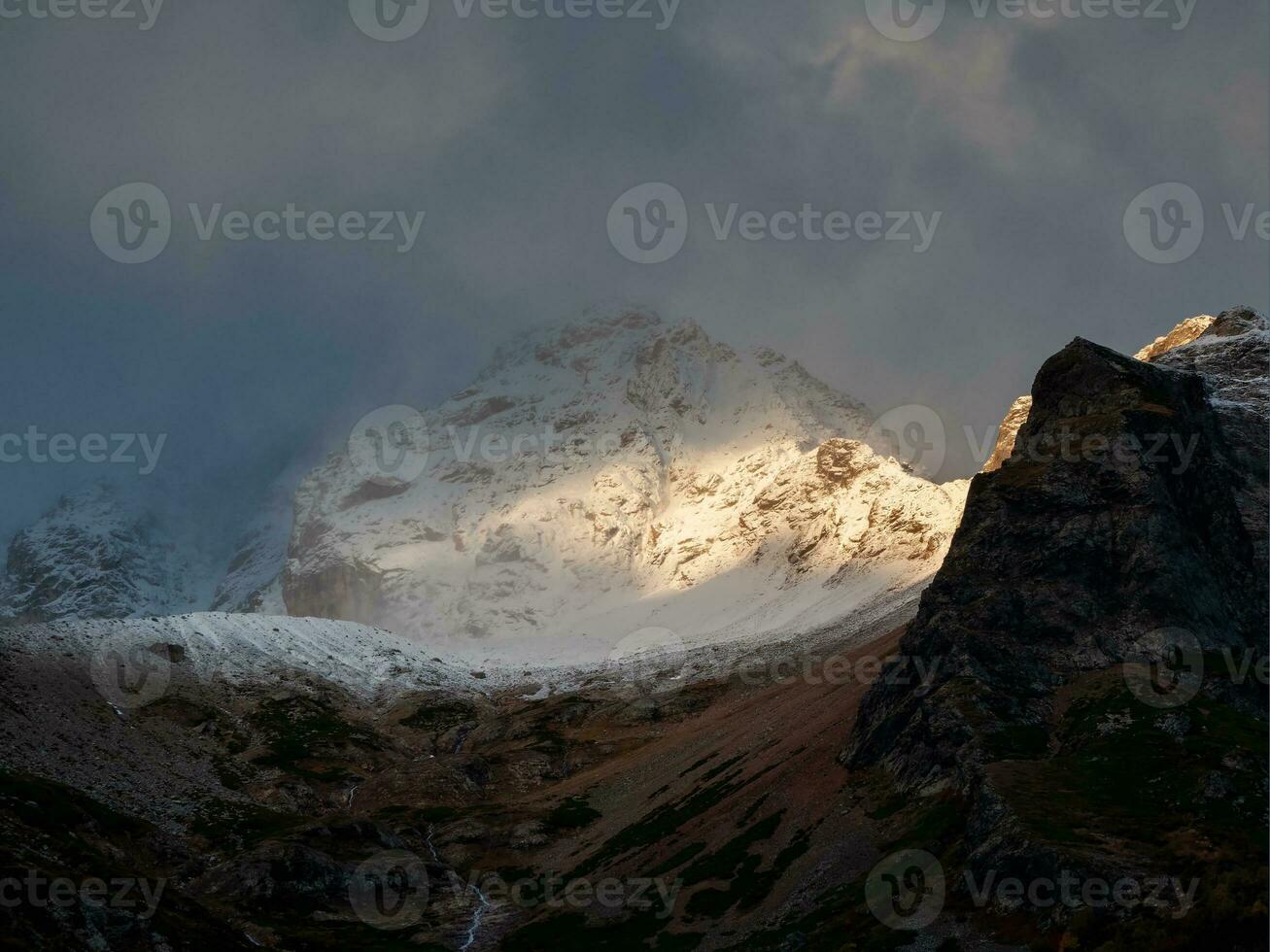 Bright ray of sunlight on a glacier. Contrasting mountains at sunrise. Morning mountain landscape with snowy rock in blizzard. Nature background of rocky mountain with sharp rock and cloudy sky. photo