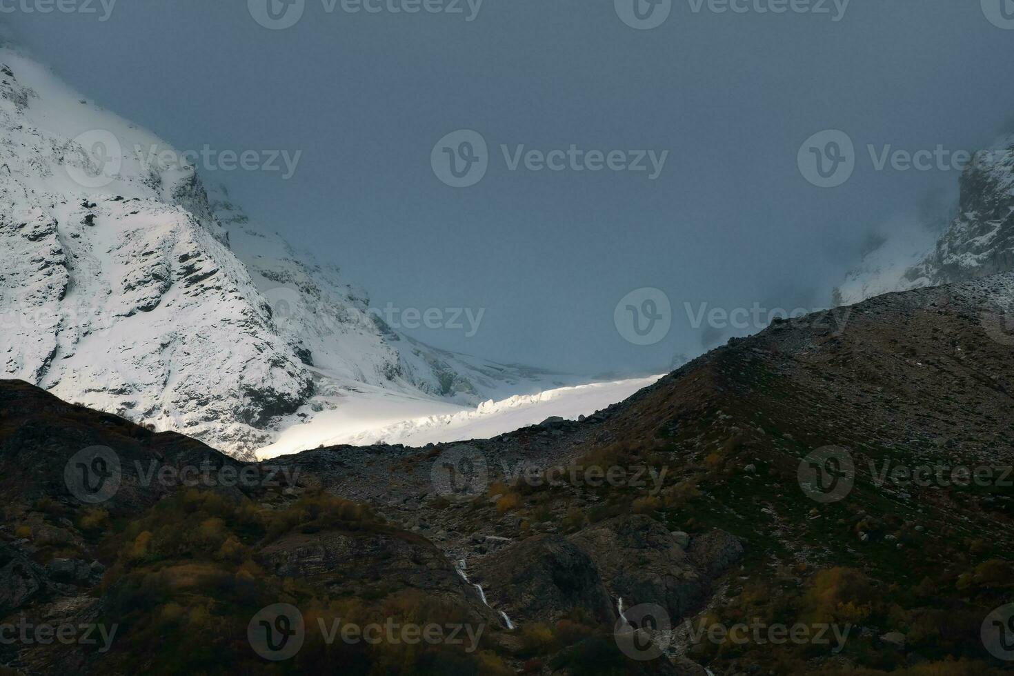 Narrow ray of sunlight on a glacier. Contrasting mountains at sunrise. Morning mountain landscape with snowy rock in blizzard. Nature background of rocky mountain with sharp rock and cloudy sky. photo