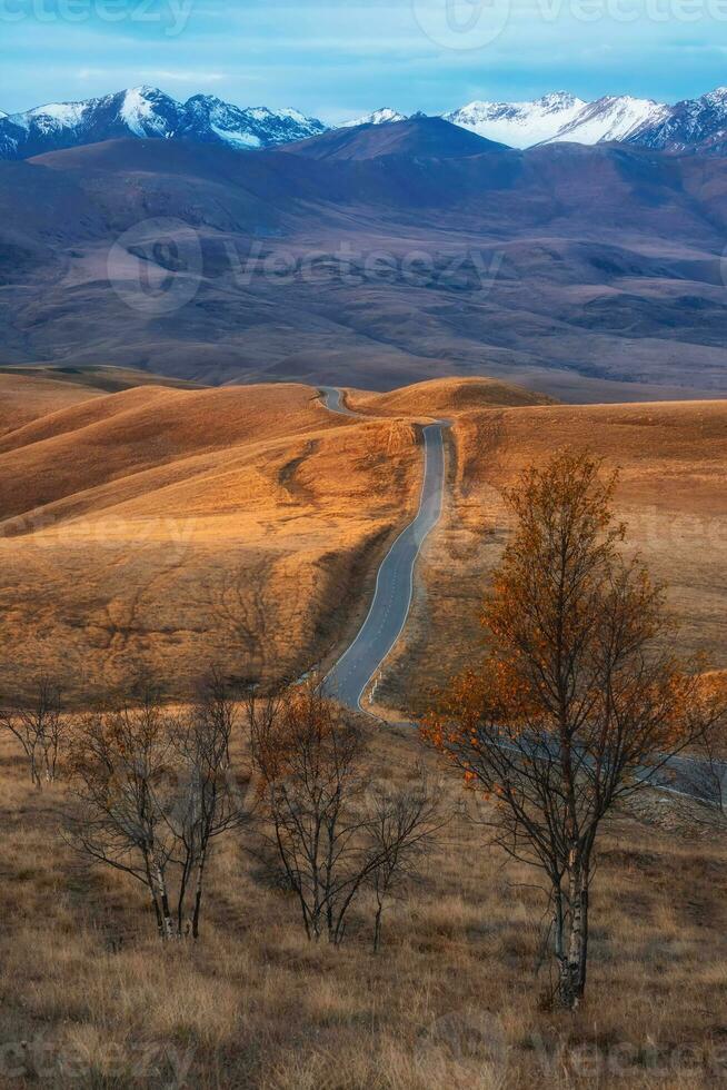 Vertical view of a road stretching into the distance through the picturesque autumn hills. Winding asphalted clean road stretches into the distance to the snow-capped mountains. photo