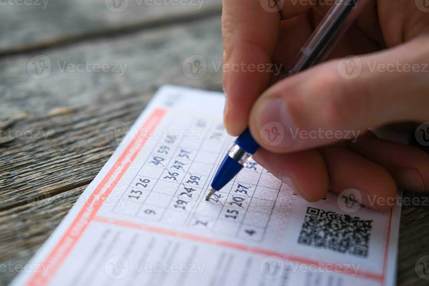 A man is hand crosses out the numbers in a lottery ticket with a pen on a wooden table. Won. Gambling addiction, addiction. photo