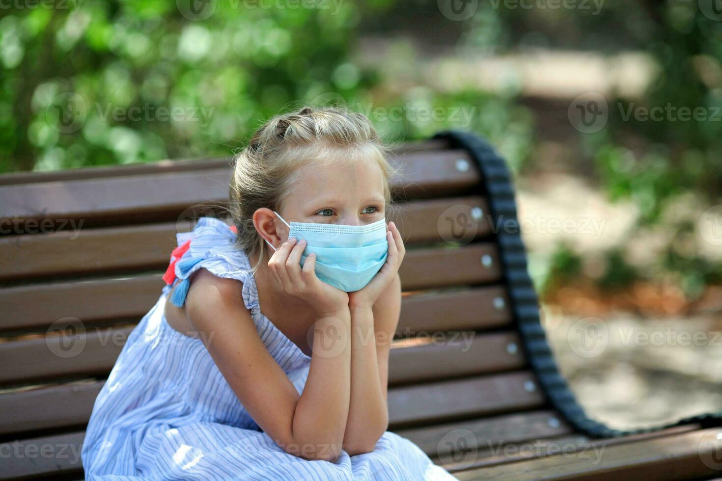 A child in a medical mask sits on a bench. A sad girl is sitting on a bench. Walk in the face of the covid-19 pandemic. photo