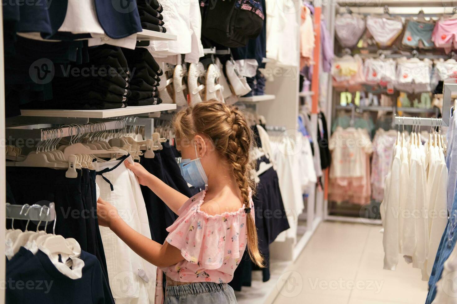 Little girl in a medical mask chooses clothes for school. School uniform store for children. Preparing for school after quarantine Covid - 19. Protective mask for the prevention of coronavirus. photo