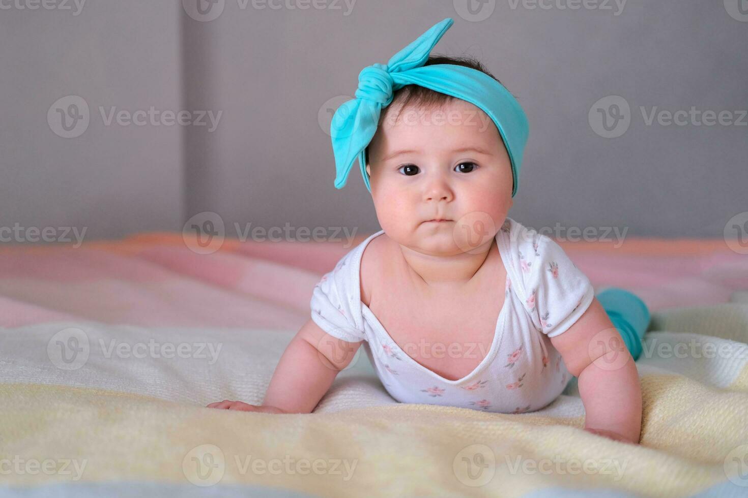 Baby at 6 months raises his body and learns to crawl. A little girl lies on her stomach on the bed and while looking at the camera. A beautiful headband for a child. photo