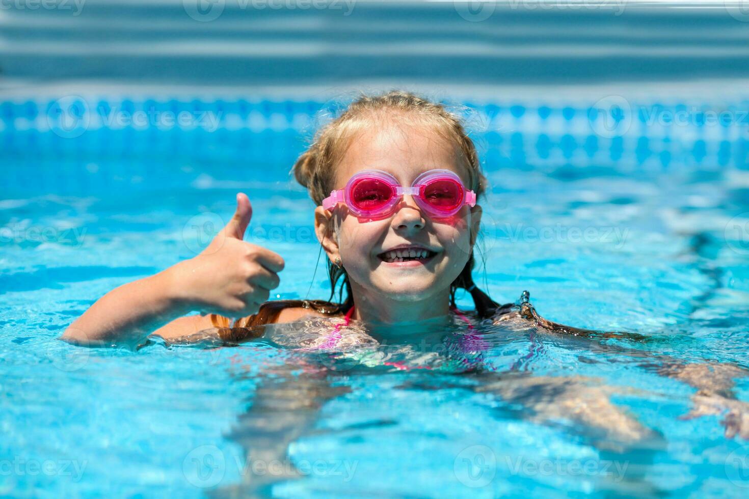 Cute little girl smiling in goggles in the pool on a sunny day. The child showing thumbs up like gesture looking camera . Summer vacation photo