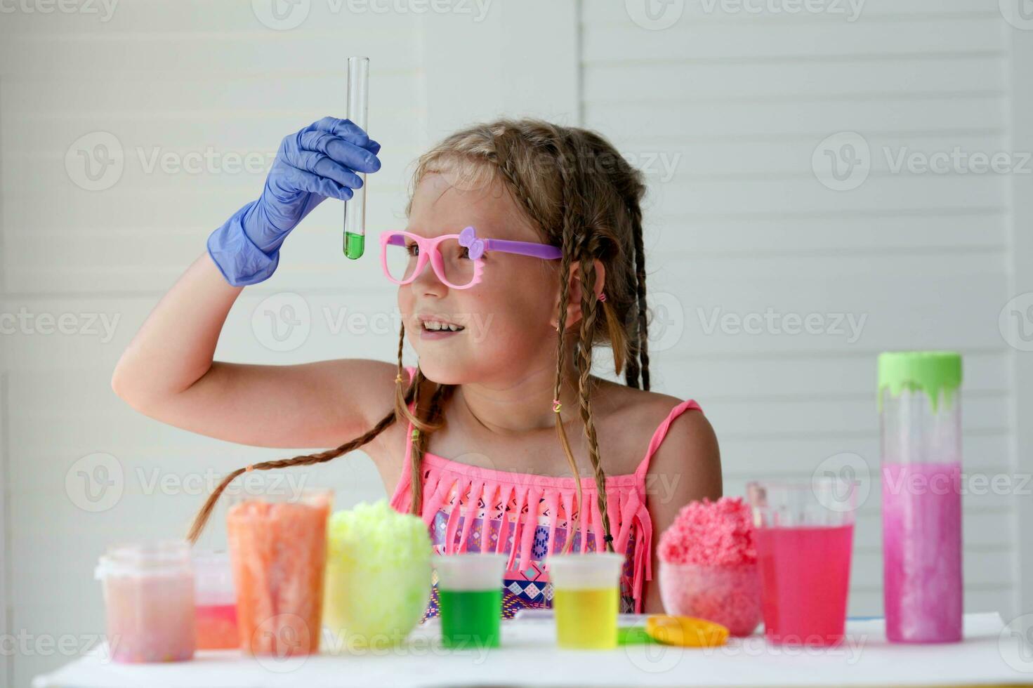 A little girl in pink glasses conducts experiments, looks at the camera and smiles. A child in rubber gloves takes a green liquid with a pipette and adds it to the test tube. Experiment. photo