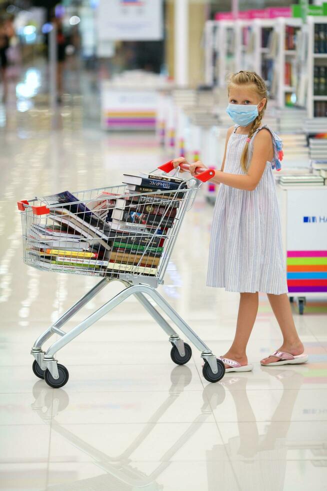 krasnodar, Rusia julio dieciséis, 2020. el niño en un médico máscara tiene lleno un cesta de libros. pequeño niña en el libro almacenar. venta. niño en el biblioteca con un carrito de libros. foto