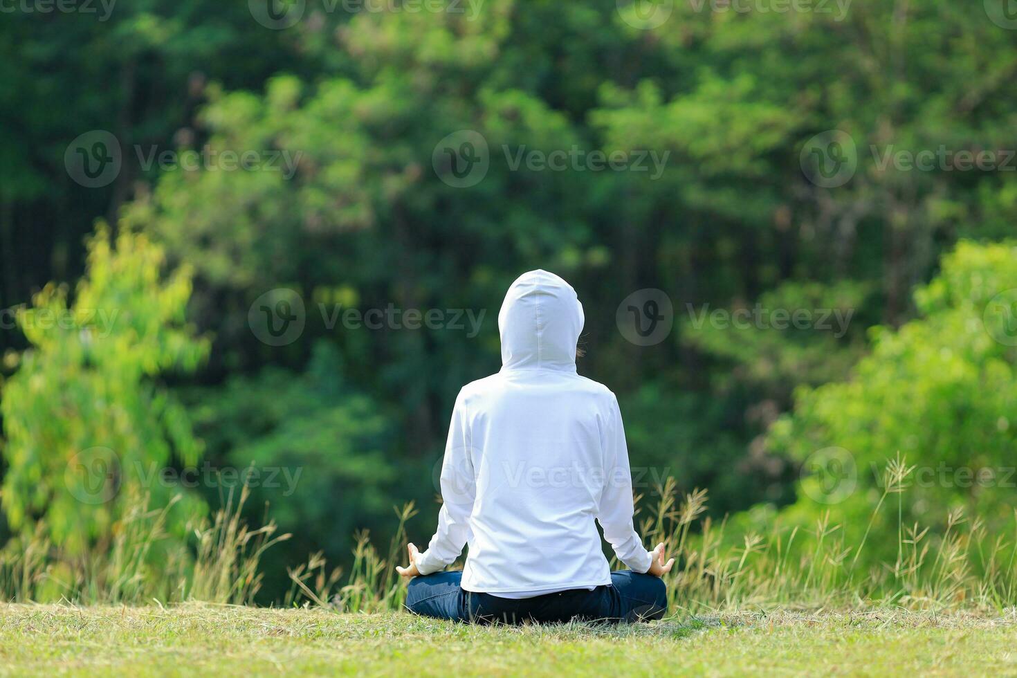 posterior ver de mujer en capucha es relajantemente practicando meditación yoga en bosque lleno de césped prado en verano a alcanzar felicidad desde interior paz sabiduría con Mañana ligero para sano mente y alma foto