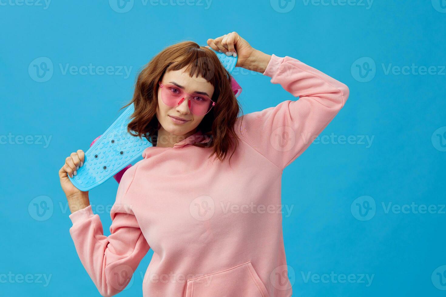 Happy cheerful pretty redhead lady in pink hoodie sunglasses with penny board posing isolated on blue studio background. Copy space Banner Offer. Fashion Cinema. Holiday activity concept photo