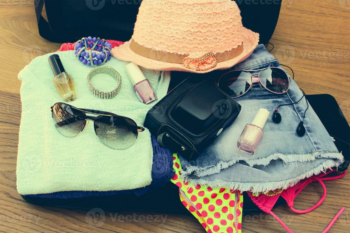 Open the suitcase with tourist things women's hat, swimsuit, camera, denim shorts, dresses, sunglasses, perfumes, nail polish, bracelets. Toned image. photo