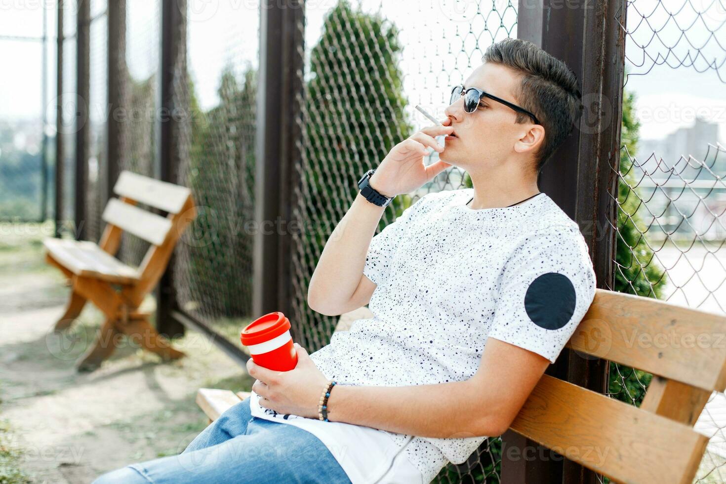 Young depressed man sitting on a bench and smokes. Guy holding a cup of coffee in his hand. photo