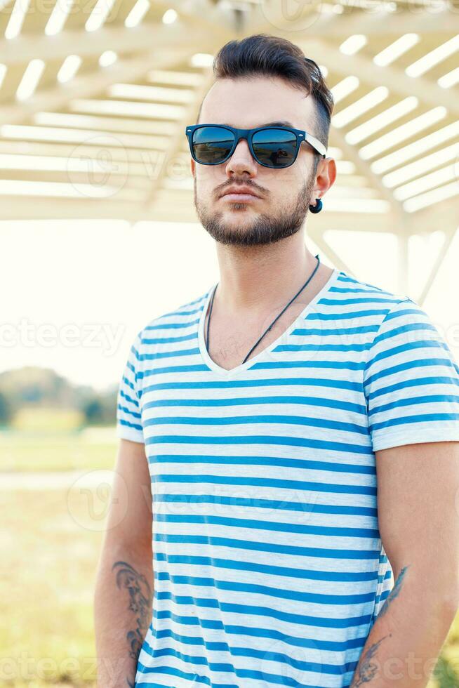 joven hermoso hipster chico con barba en un elegante camiseta y Gafas de sol en un soleado verano día a el playa. foto