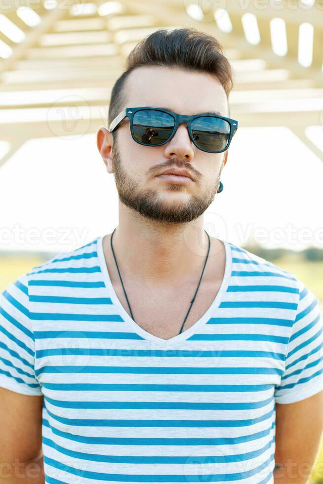 Handsome man in a t-shirt with the beard in sunglasses standing on the beach. photo