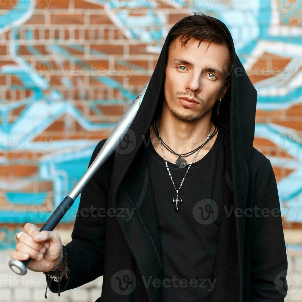 Handsome man with a bat in black clothes in the hood near the brick wall with graffiti photo