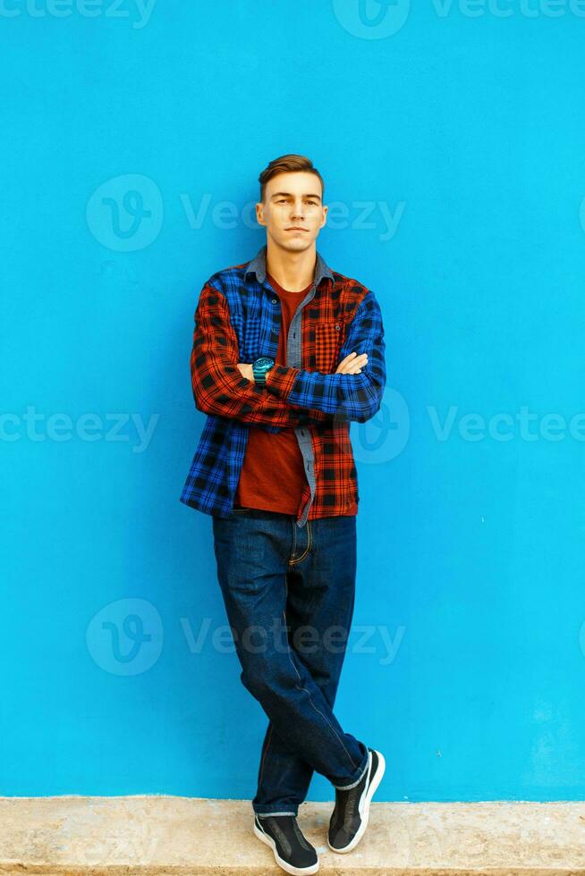 Stylish handsome man in a fashionable shirt, sneakers and jeans stands near a bright blue wall. photo