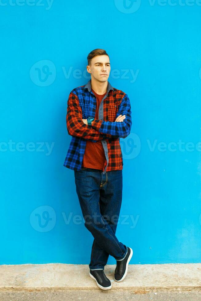 Young handsome man in stylish clothes and sneakers poses near the blue wall photo