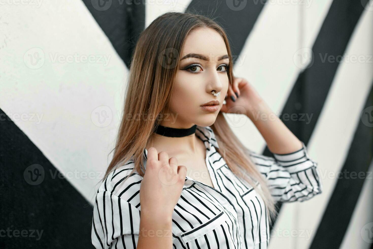 bonito mujer en un blanco blusa con negro rayas cerca el pared con líneas. arete en el nariz foto