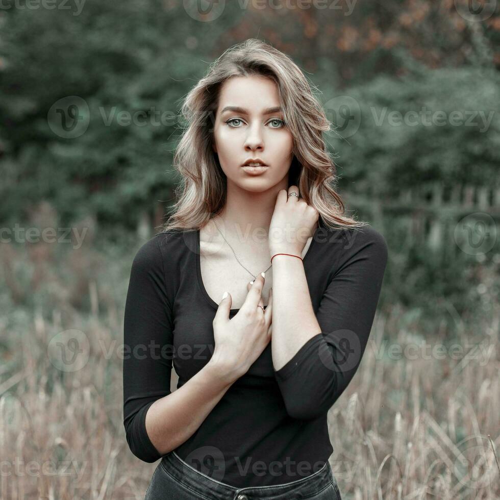 hermosa joven rubia niña en un negro camiseta en un campo cerca arboles foto