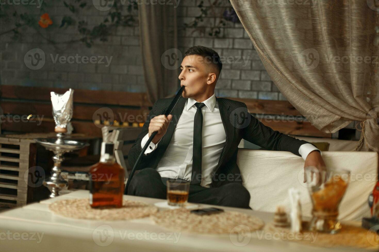 joven elegante hombre en un traje descansa en el bar. hombre Bebiendo whisky y de fumar un narguile. foto