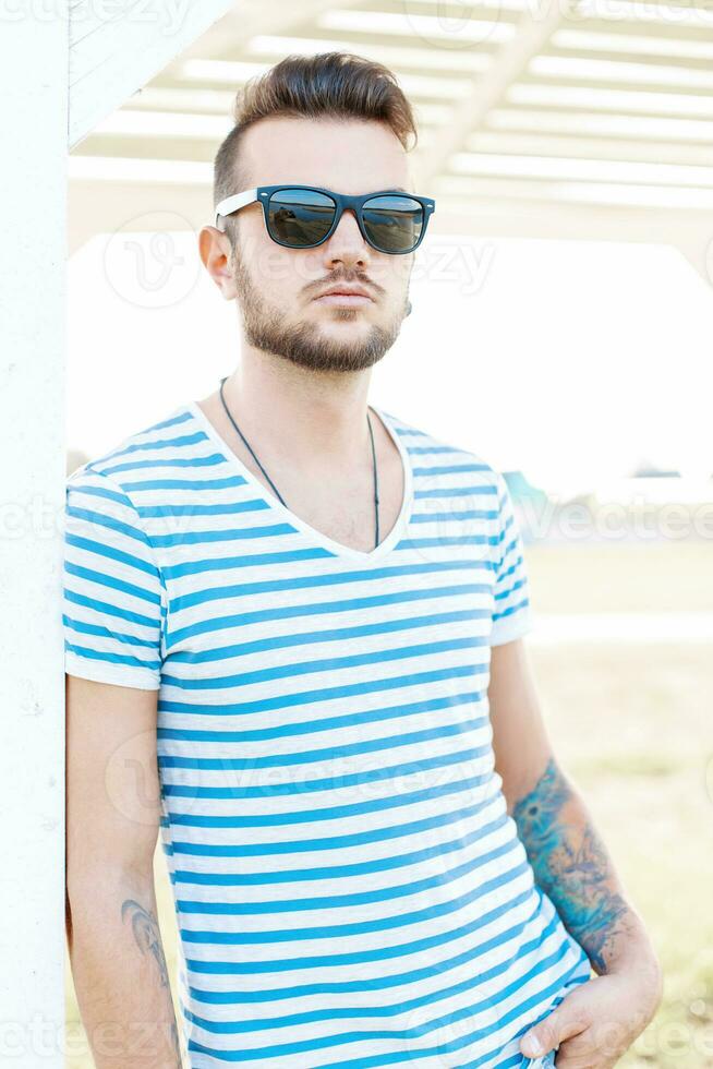 Handsome hipster man with a beard in sunglasses standing on the beach on a sunny day. photo