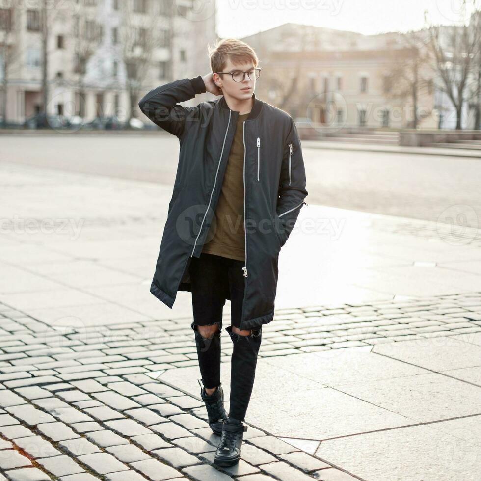 Handsome young man with glasses in a black jacket in torn jeans and black shoes goes through the city photo
