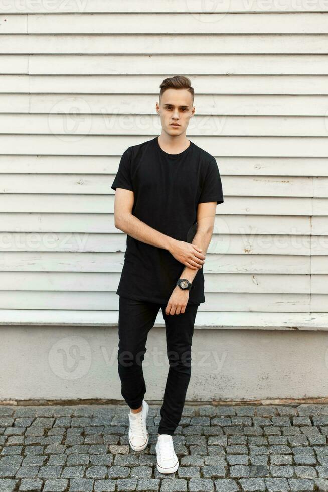 Young handsome man in black stylish dress and white sneakers posing near a light wooden wall. photo