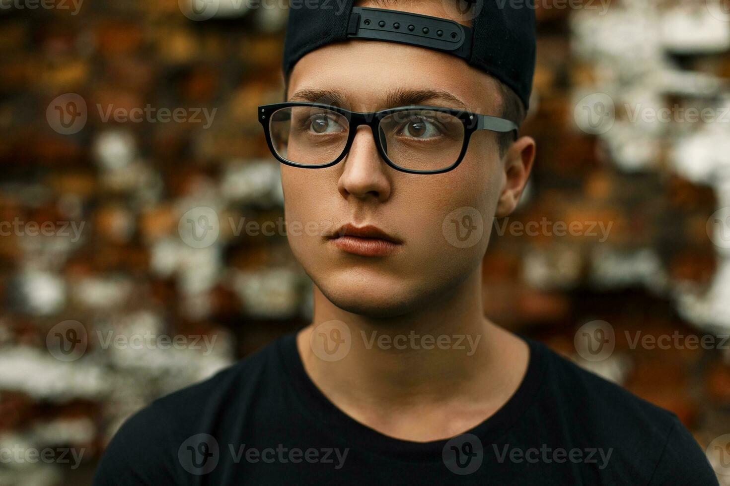 Young stylish man with glasses and a black cap stands near a red brick wall photo