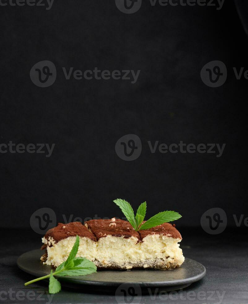 A piece of tiramisu sprinkled with cocoa, on top of a sprig of fresh mint on a black background photo